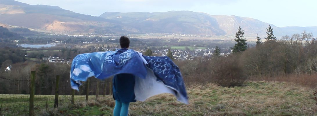 A person stands on a hillside, with their outstretched arms covered in patterned blue blankets that blow in the wind.