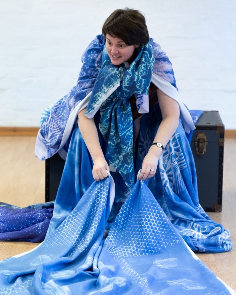 Claire holds a blanket low over the ground in front of her, whilst covered in blankets and sat on a blue vintage chest.