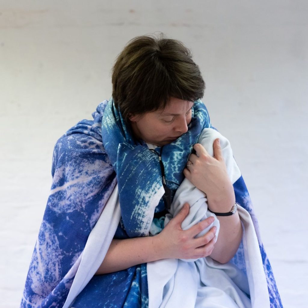 Claire cradles a white blanket as though it is a small baby. She has blue blankets over her shoulders and around her neck.