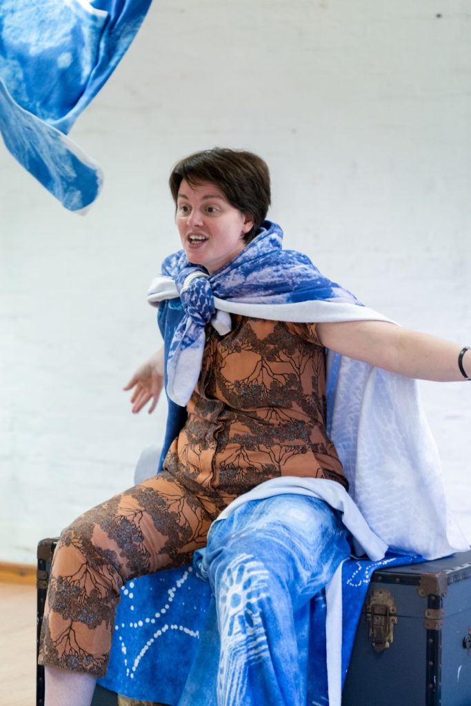 Claire, w white woman with short brown hair is dressed in a brown jumpsuit, with blue blankets over her shoulders and leg. One blanket flies into the air as she spreads her arms wide.